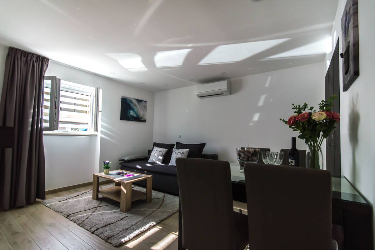 A modern living room with a sofa, coffee table, and chairs. There is a window with blinds, a painting on the wall, and a vase with flowers on a table.