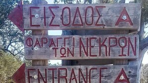 Un letrero de madera con indicaciones y nombres, incluyendo 'EIZOAOZ A', 'ΦΑΡΑΤΕΝ ΝΕΚΡΟΝ', 'ENTRADA' y 'DESFILADERO DE LOS MUERTOS'.