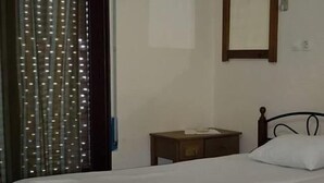 Una cama individual con estructura de madera y ropa de cama blanca, una mesita de noche pequeña, un espejo fijado a la pared y una ventana con persianas verticales.
