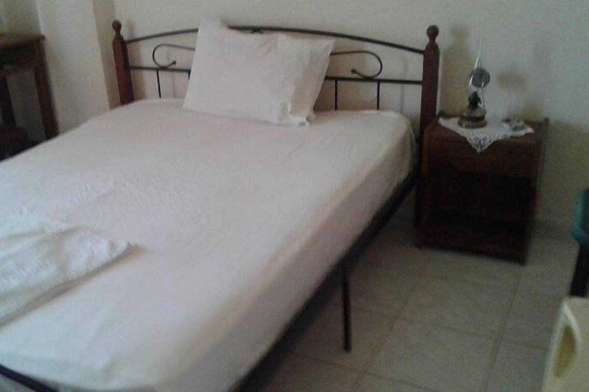 A single bed with a white mattress and a wooden headboard. There is a small bedside table with a lamp and a decorative item on it. The room has a tiled floor and a plain wall.