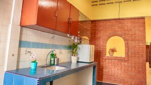 Une cuisine avec un plan de travail bleu, un évier blanc et des placards rouges. On aperçoit une plante en pot sur le plan de travail et un mur de briques avec une petite fenêtre.