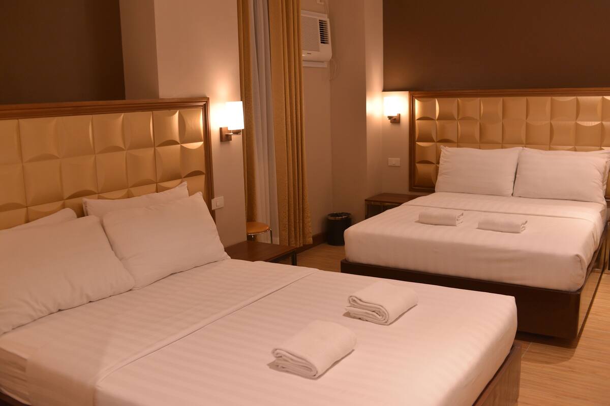 Habitación de hotel con dos camas, cada una con toallas ordenadas, cabeceros de madera y lámparas fijadas a la pared.
