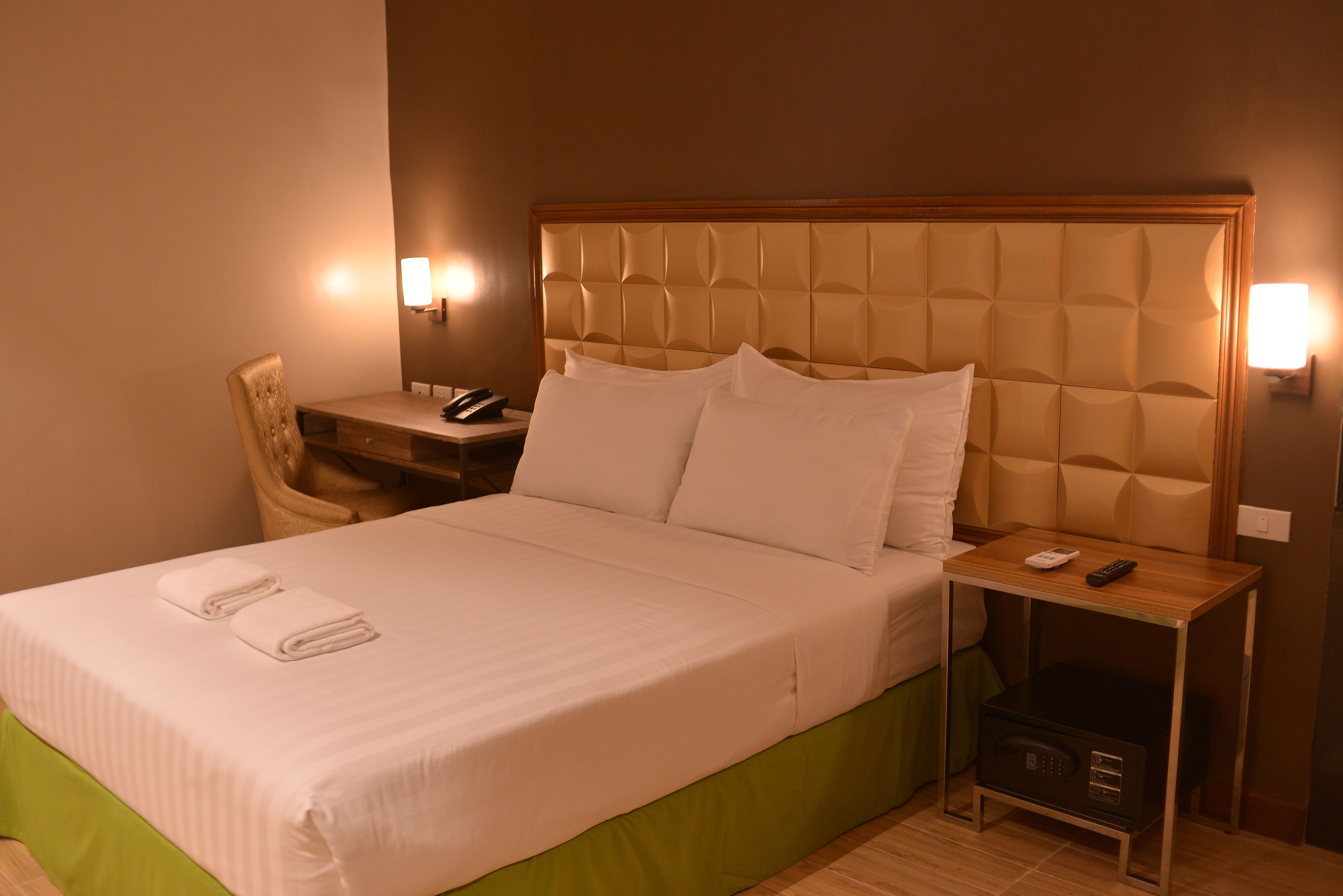 A neatly made bed with a green bed skirt, a bedside table with a lamp, a chair, and a small bedside table with a remote control.