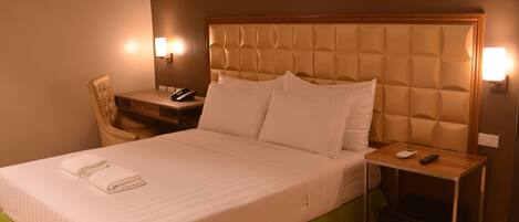 A neatly made bed with a green bed skirt, a bedside table with a lamp, a chair, and a small bedside table with a remote control.