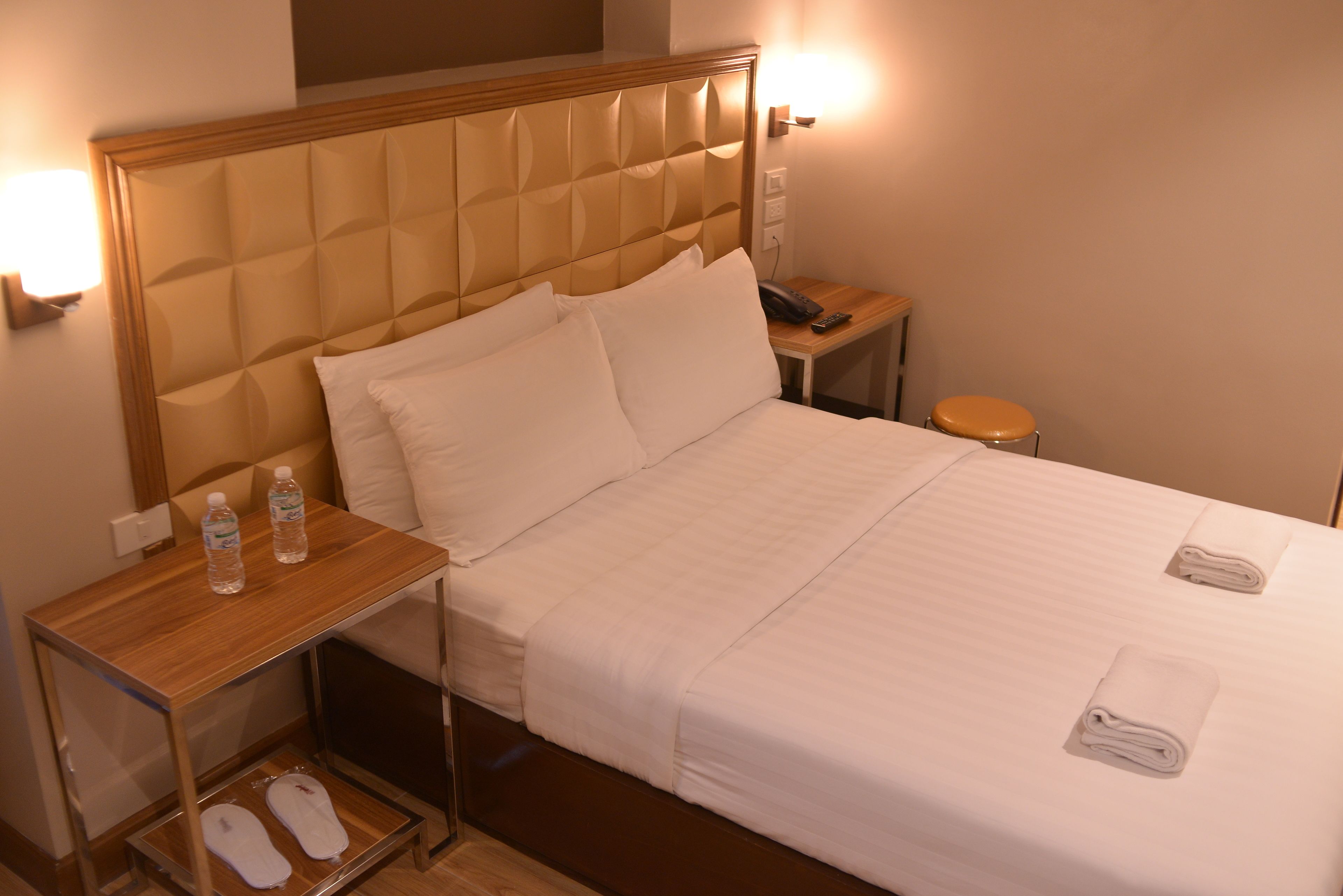 A hotel room with a large bed, a bedside table with a phone and water bottles, and a small stool.