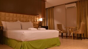 Een hotelkamer met een groot bed, een groene rok, twee stoelen, een kleine tafel en een airconditioner.