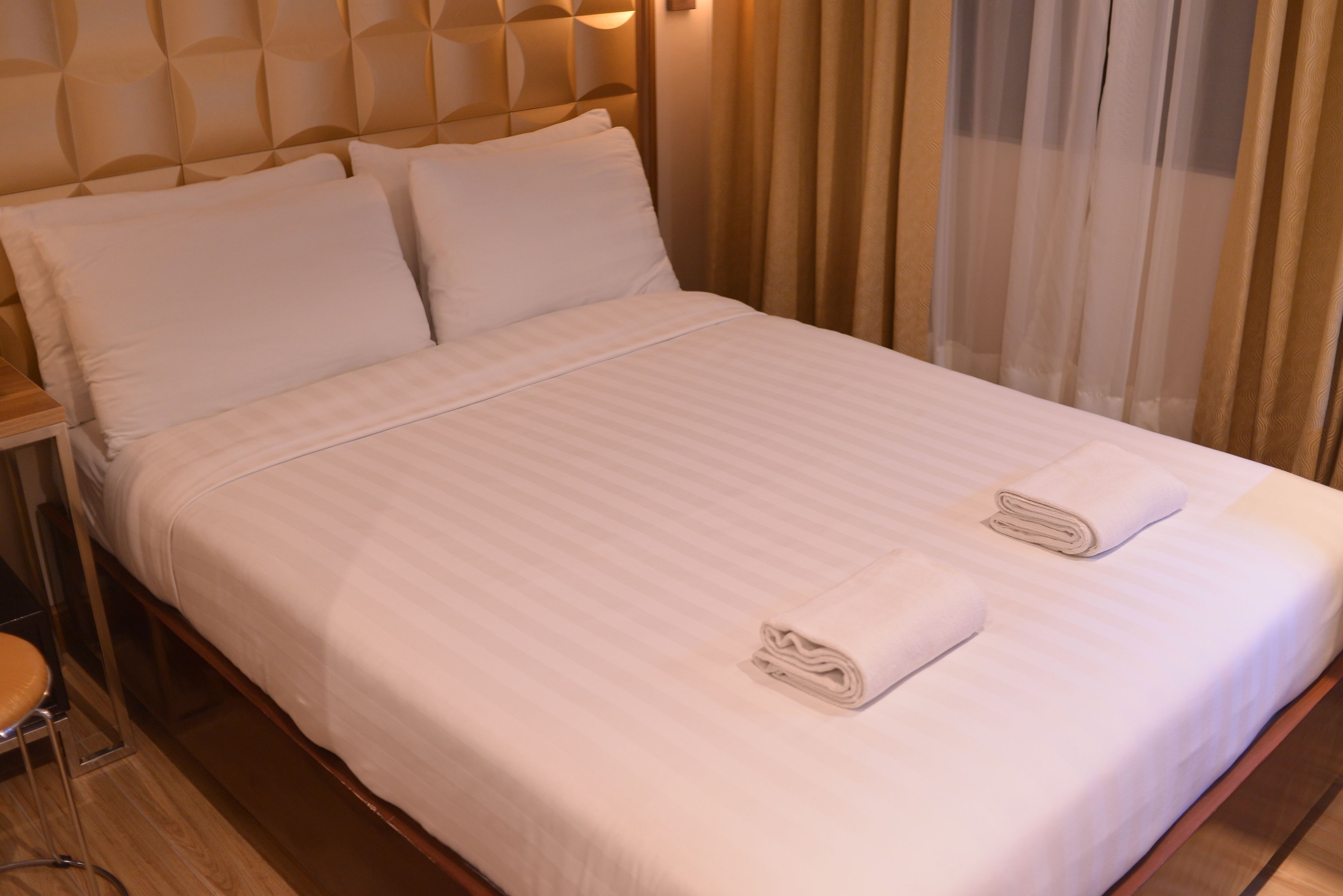 A neatly made bed with white linens and two folded towels, a wooden headboard, and a wooden nightstand with a chair.