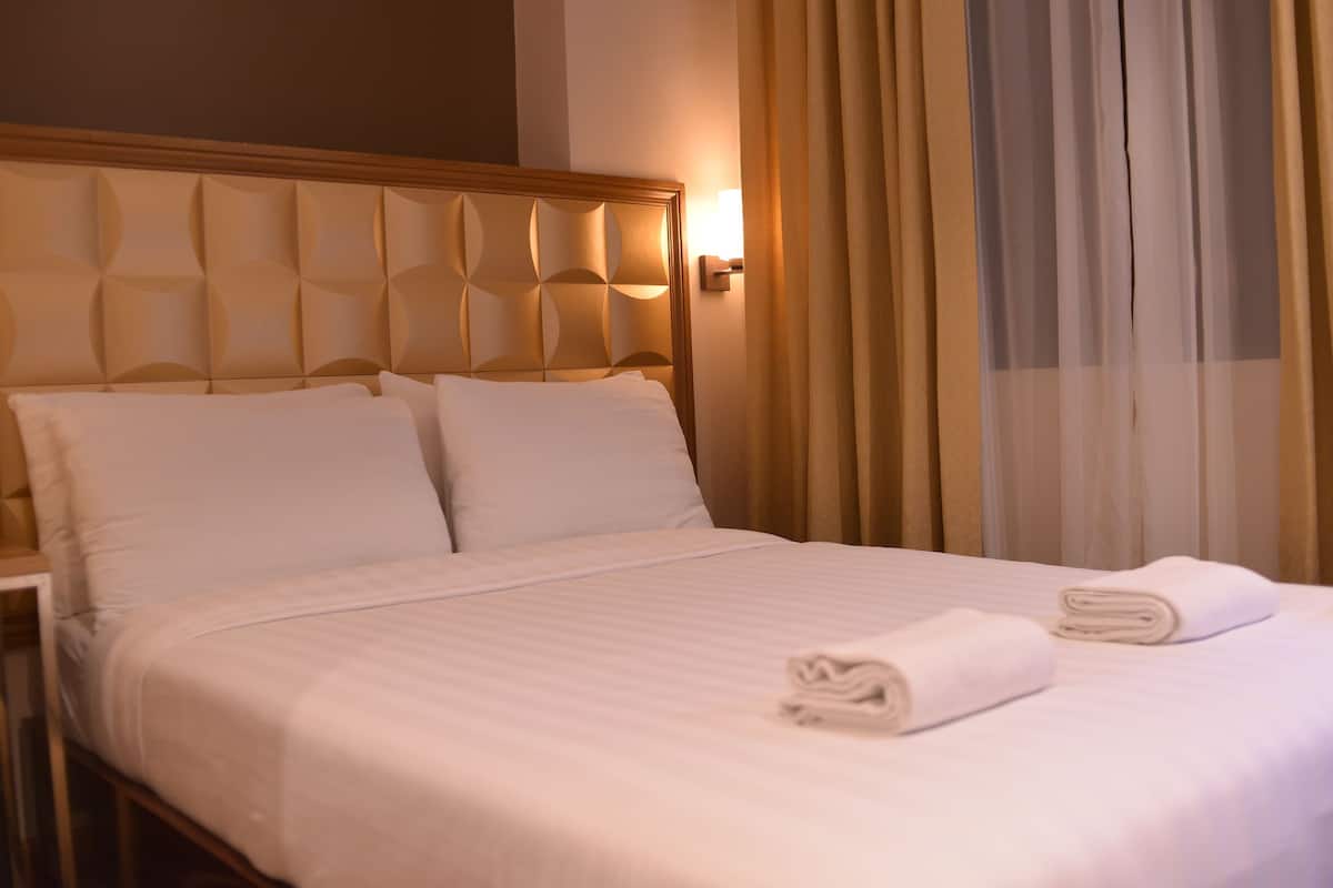Una cama bien tendida con sábanas blancas y dos toallas enrolladas, un cabecero de madera y una lámpara de pared.