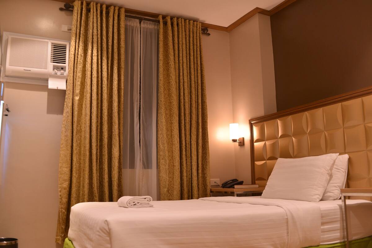 Habitación de hotel con cama, mesita de noche con teléfono, aire acondicionado de pared y ventana con cortinas.