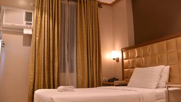 A hotel room with a bed, a bedside table with a phone, a wall-mounted air conditioner, and a window with curtains.