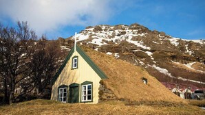 Ein kleines, eingeschossiges Haus mit steiler Dachneigung, auf einem grasbewachsen Hang gelegen, mit einem Berg im Hintergrund.