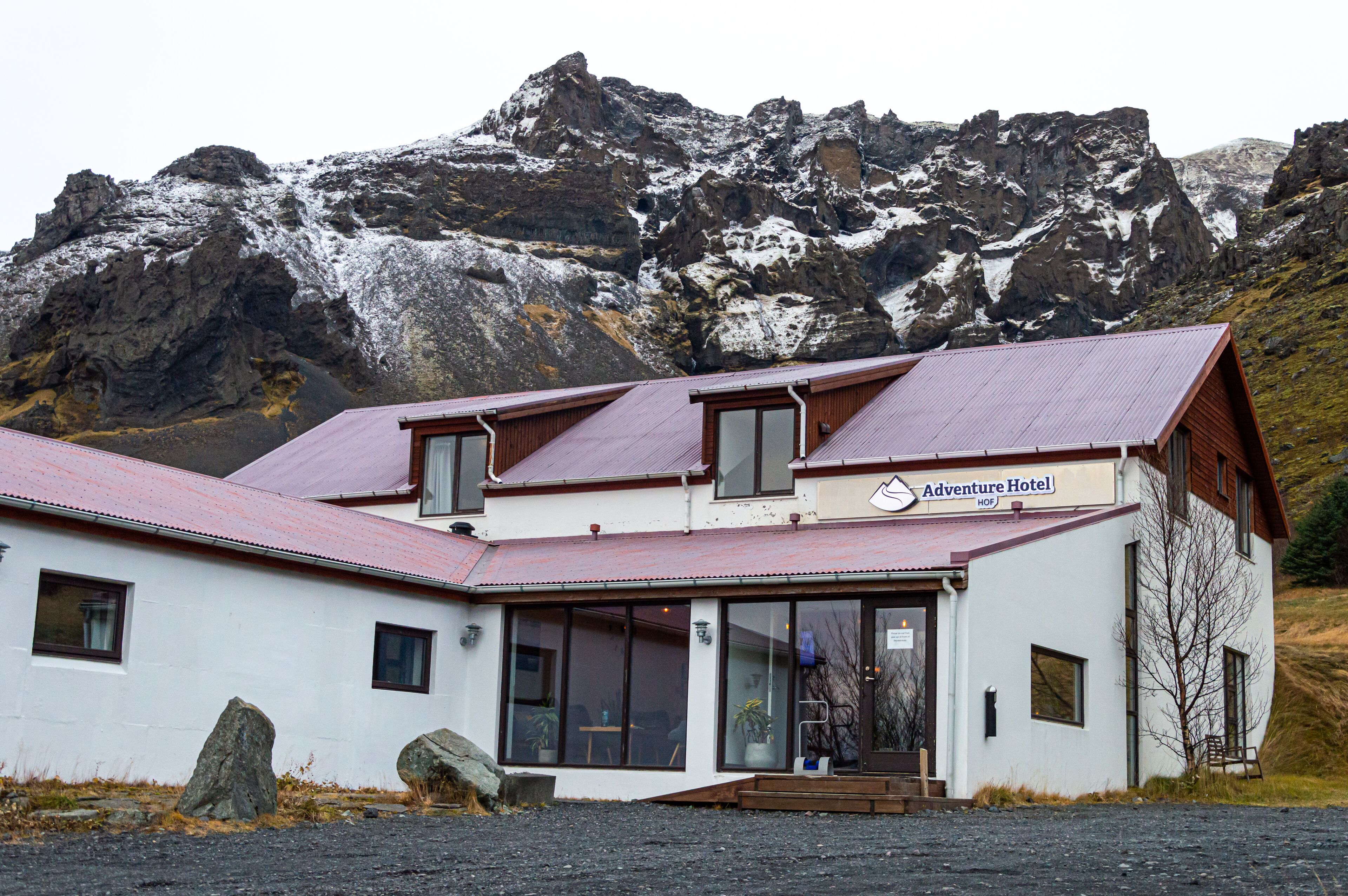 A white building with a red roof, labeled "Adventure Hotel." It features large windows, a wooden staircase, and is surrounded by rocky mountains with snow.