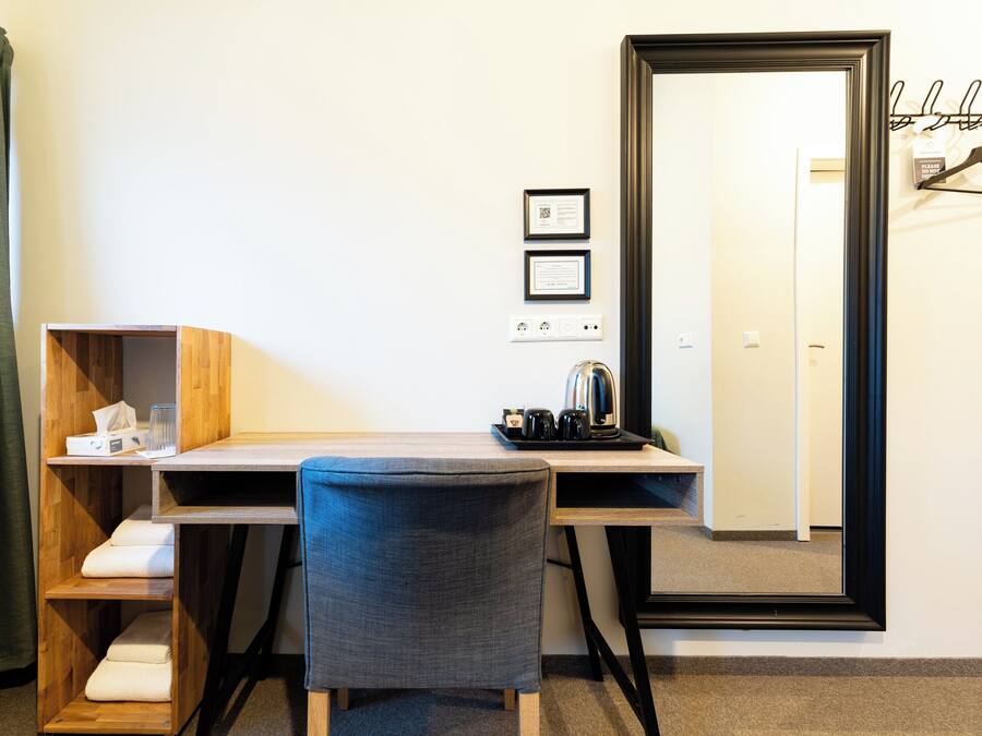 Quarto de hotel com escrivaninha, cadeira e um espelho grande. Há dois certificados emoldurados na parede, uma pequena prateleira com lenços de papel e um copo, e uma bandeja com chaleira e xícaras.