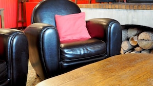 Un fauteuil en cuir avec un coussin rouge, une table en bois et une cheminée où sont empilés des bûches.