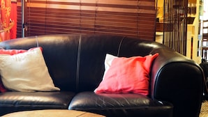 Un canapé en cuir noir avec deux coussins, un blanc et un rouge, et une table en bois dans une pièce dotée de stores en bois et d’un sol carrelé.