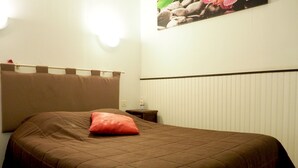 Un lit bien fait, avec une couverture marron et un seul oreiller rouge, une table de chevet avec une bouteille, et un luminaire fixé au mur.