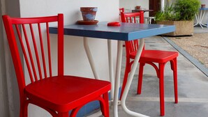 Un espace extérieur aménagé avec des sièges rouges et bleus, comprenant une table et des chaises, une plante en pot et un mur blanc en arrière-plan.