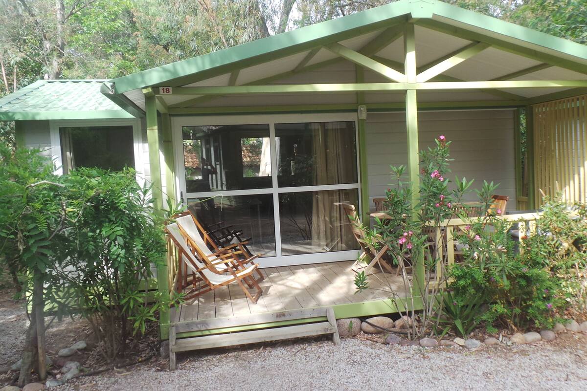 A covered outdoor seating area with a table and chairs, surrounded by greenery and a gravel path.