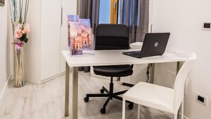 Espaço de escritório moderno com uma secretária branca, cadeira de escritório preta e um laptop. Há um vaso com flores à esquerda e uma moldura com fotografia na secretária.