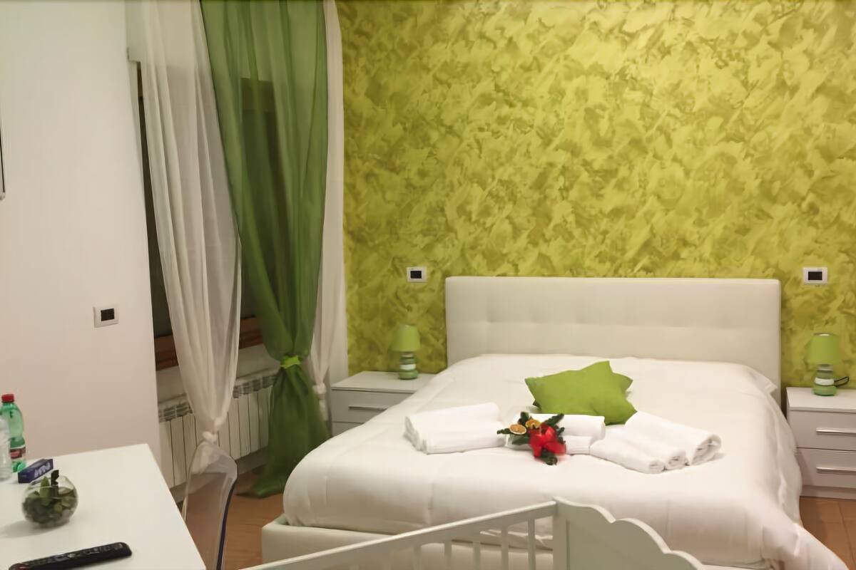 Une chambre à coucher bien rangée, avec un lit, des tables de chevet, une petite table avec une télécommande et un rideau vert.