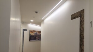 Corredor com paredes brancas, uma câmera de segurança instalada no teto, iluminação embutida e uma porta com o símbolo de número.