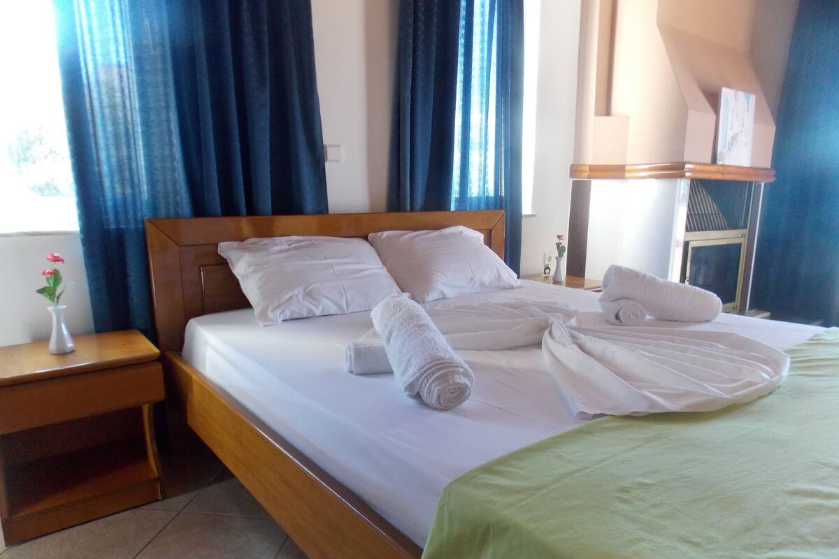 A neatly made bed with white linens and pillows, a wooden headboard, a green throw blanket, and a small vase with flowers on a nightstand.