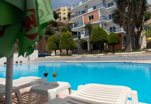 A swimming pool with lounge chairs and two glasses of wine, surrounded by buildings and trees. - Hotel Dea (Sarandë)