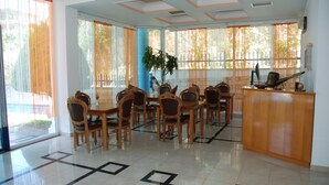 A spacious dining area with a long table and multiple chairs, large windows with curtains, and a wooden reception desk.