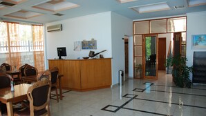 A hotel lobby with a reception desk, wooden chairs, a tiled floor, and a large window with curtains.