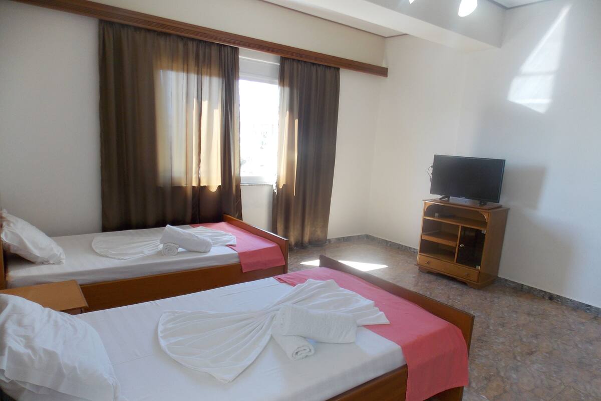 A hotel room with two beds, a television on a wooden stand, and a window with curtains.