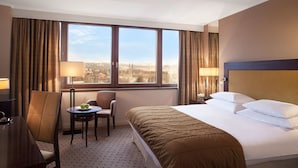 A hotel room with a large bed, a desk with a chair, a small table with a lamp, and a view of a cityscape through large windows. - Tribecca Residency (Ahmedabad)