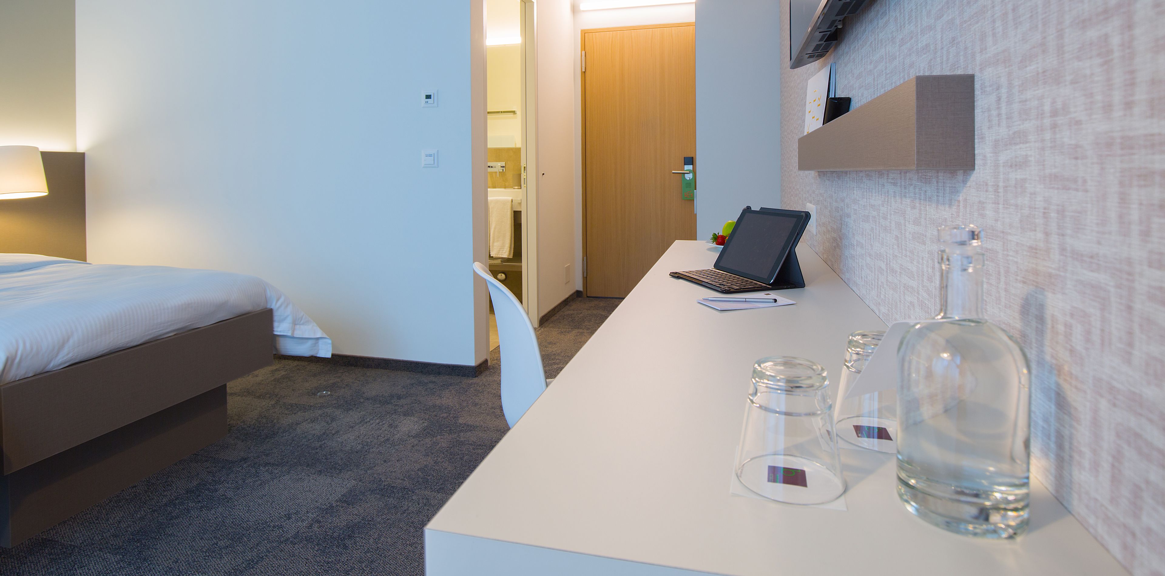 A hotel room with a bed, a desk with a laptop, a bottle, and glasses.
