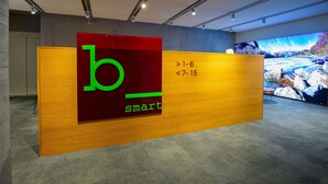 A reception desk with a large 'b smart' logo, a digital display with a scenic image, and a carpeted floor. - b smart motel Landquart (Landquart)