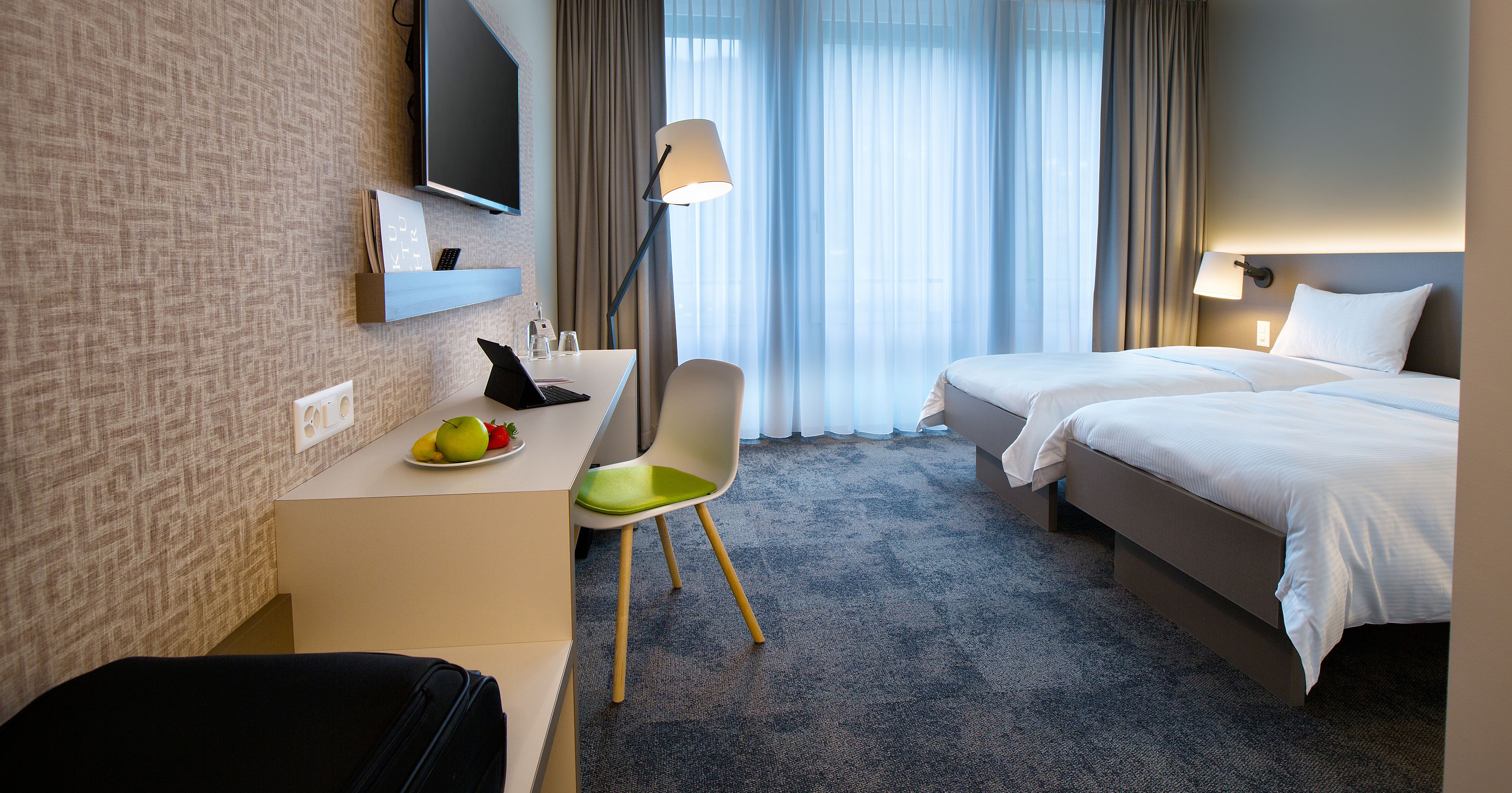 A hotel room with a desk, chair, two beds, a TV, and a window with curtains.