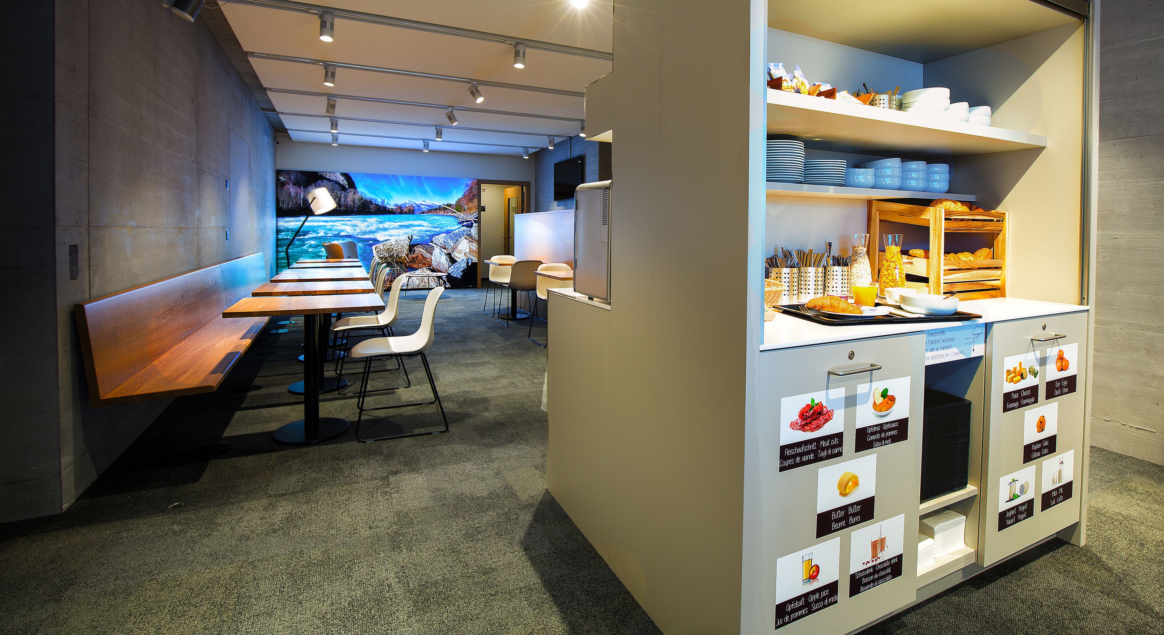 Un comedor moderno con una larga mesa de madera y sillas blancas, un área de cocina con alimentos identificados y una pantalla grande montada en la pared que muestra una imagen panorámica.