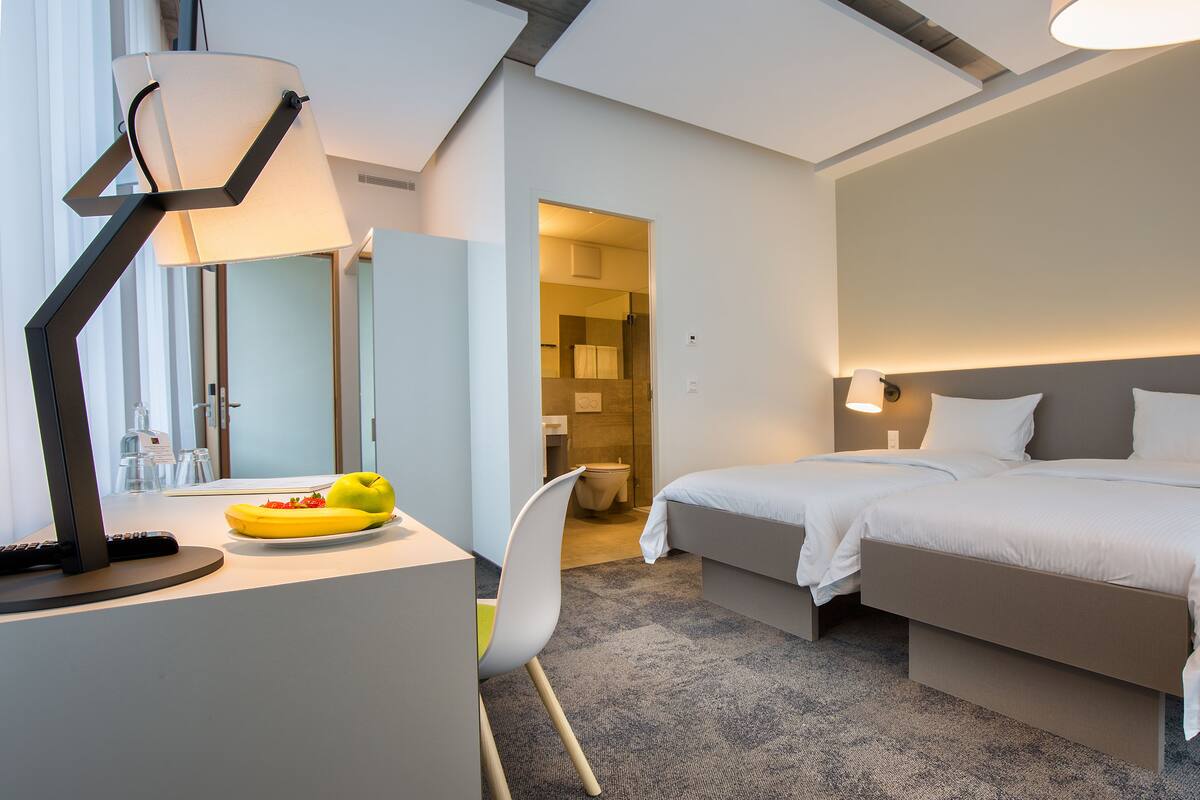 A hotel room with two beds, a desk with a lamp, a chair, and a bathroom visible through an open door.