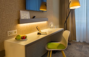 Habitación de hotel con un escritorio, una silla, una computadora portátil, un plato de fruta, un control remoto, una lámpara y un televisor montado en la pared.