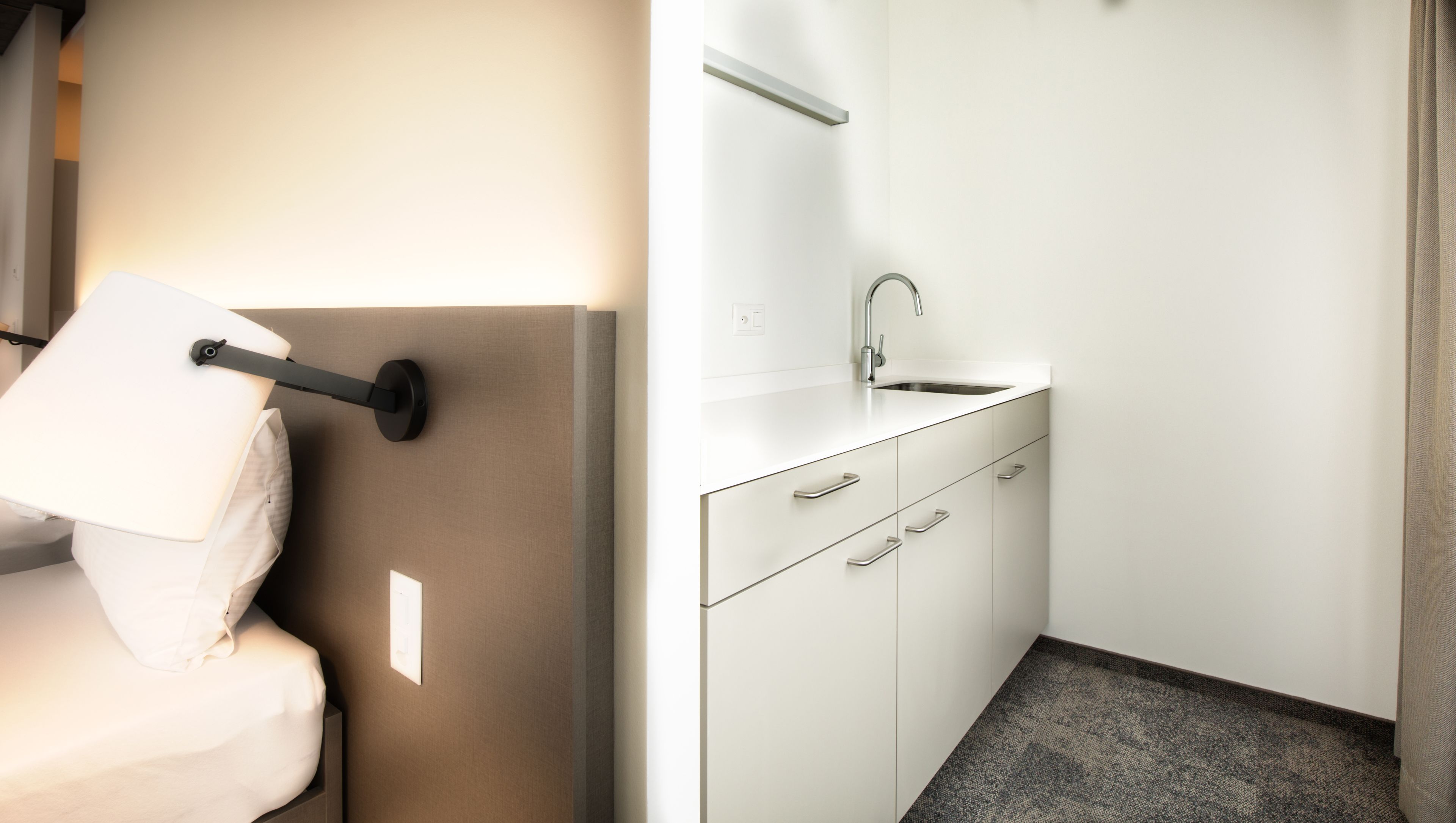 Habitación de hotel pequeña con cama, mesita de noche con lámpara, baño compacto con lavamanos y espejo, y suelo alfombrado.