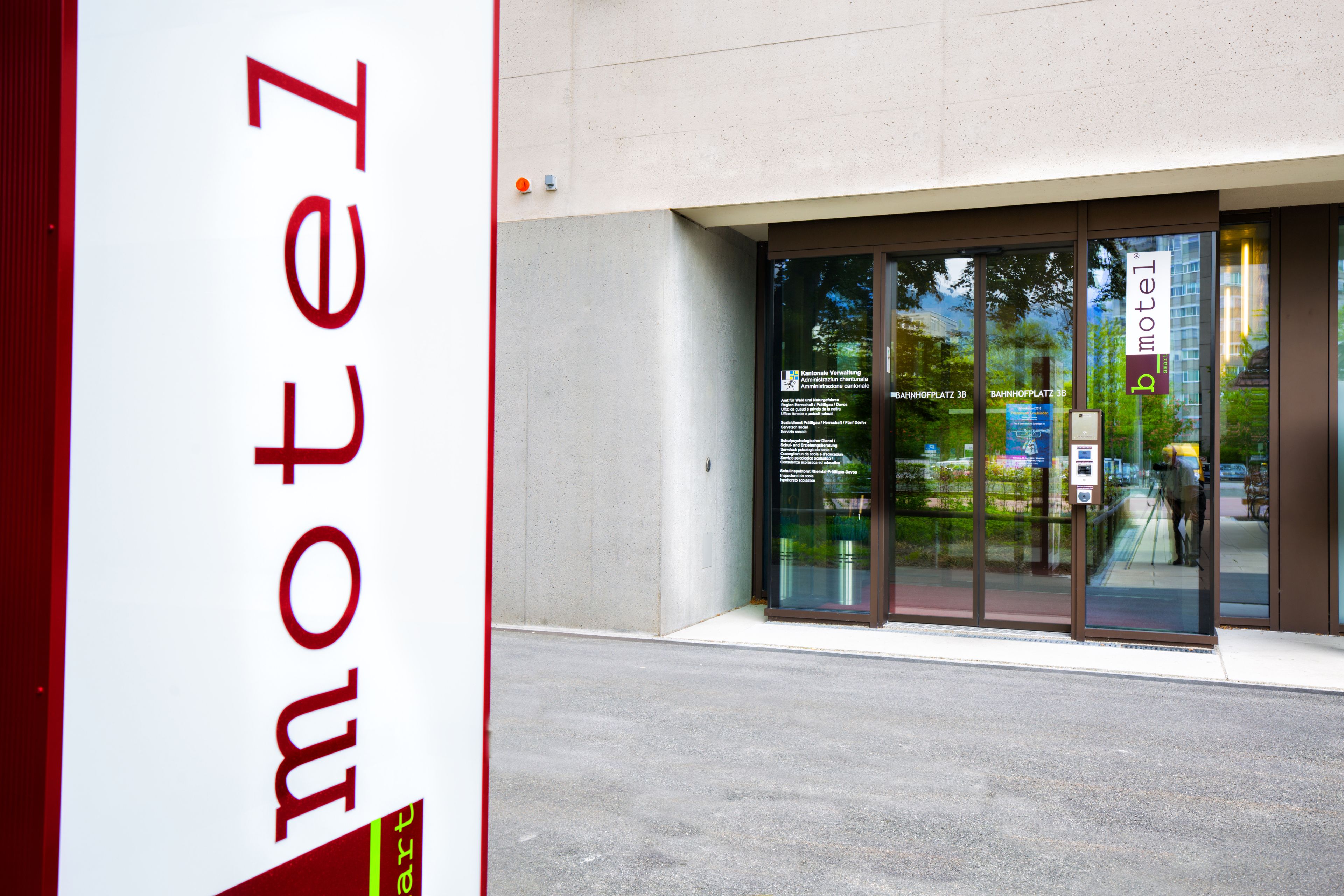 A modern building with a large glass entrance, a sign reading "motel", and a visible outdoor area with greenery.