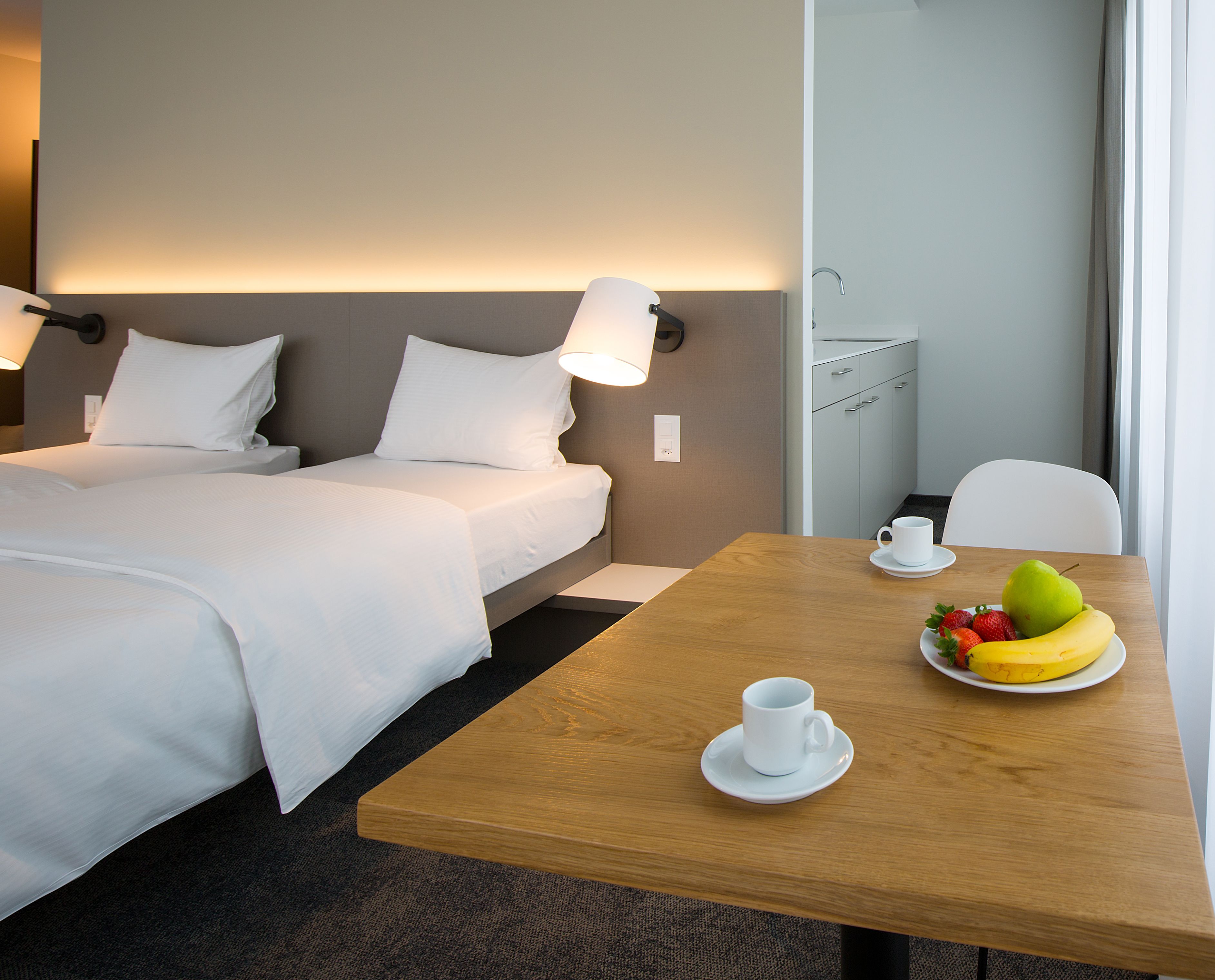 A hotel room with two beds, a wooden table with a fruit plate, a cup, and a lamp.