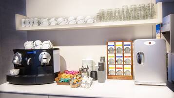 Une cuisine équipée d’une machine à café, d’un réfrigérateur, et d’une étagère où sont exposées des tasses et des soucoupes à café.