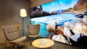 A room with two armchairs, a small round table with magazines, a floor lamp, and a large TV displaying a river scene. - b smart motel Landquart (Landquart)