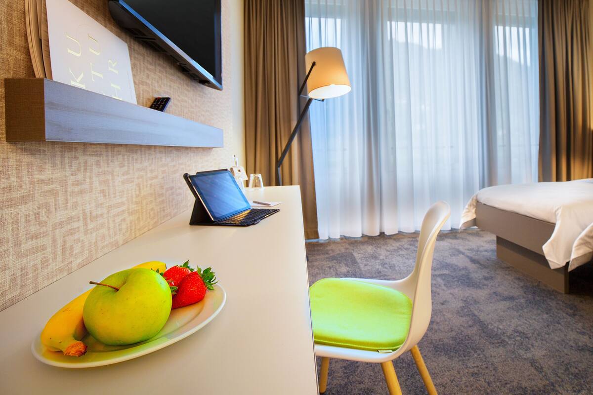 A hotel room with a bed, a desk with a laptop, a chair, a plate with fruit, and a TV mounted on the wall.