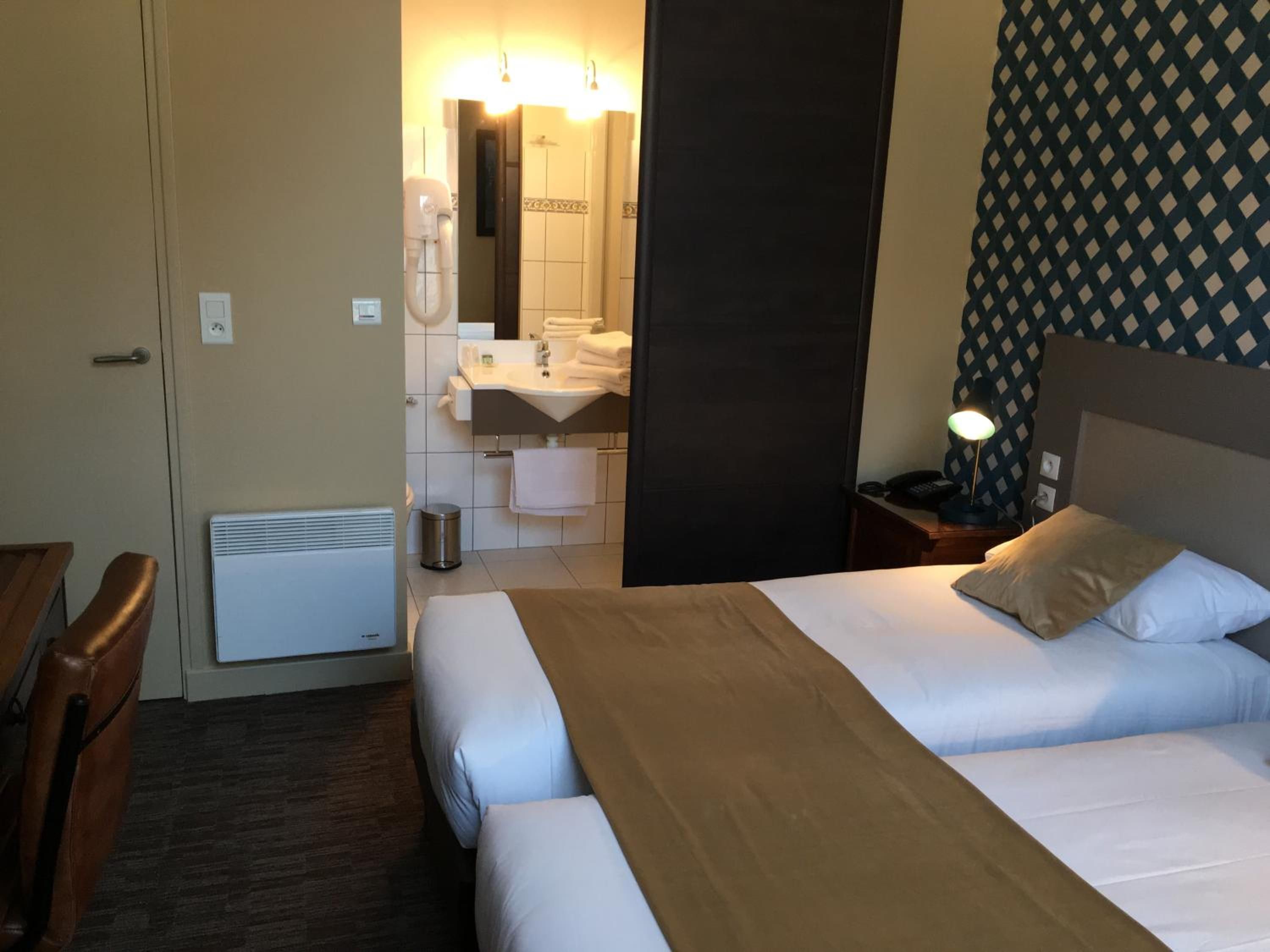 Ein Hotelzimmer mit Bett, Nachttisch, Lampe und einem Badezimmer mit Waschbecken und Spiegel.