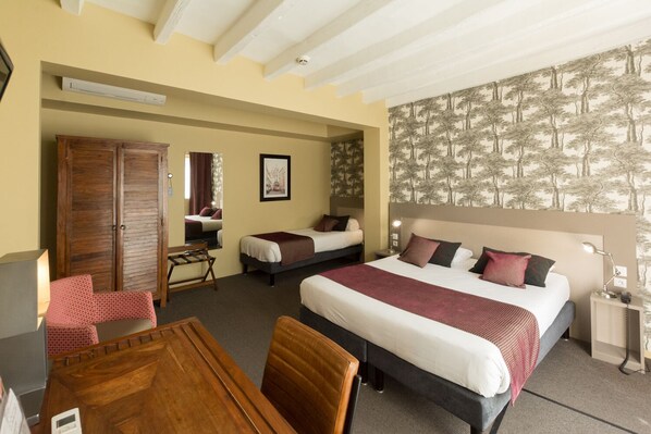 A hotel room with a large bed, two smaller beds, a wooden wardrobe, a desk with a chair, and a mirror.