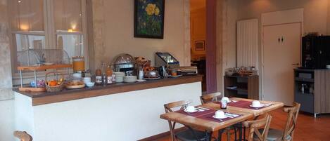 Una barra de desayuno con diversos alimentos y bebidas, una mesa de comedor con sillas y una zona de cocina al fondo.
