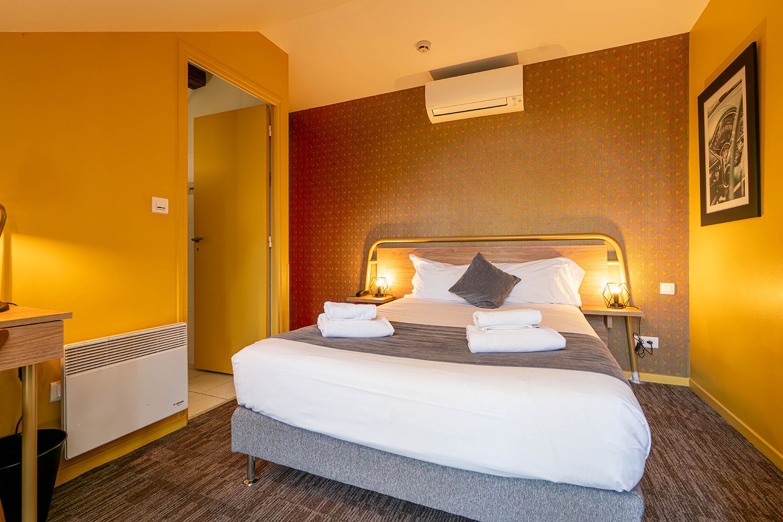 A hotel room with a bed, bedside tables, an air conditioning unit, and a framed picture on the wall.