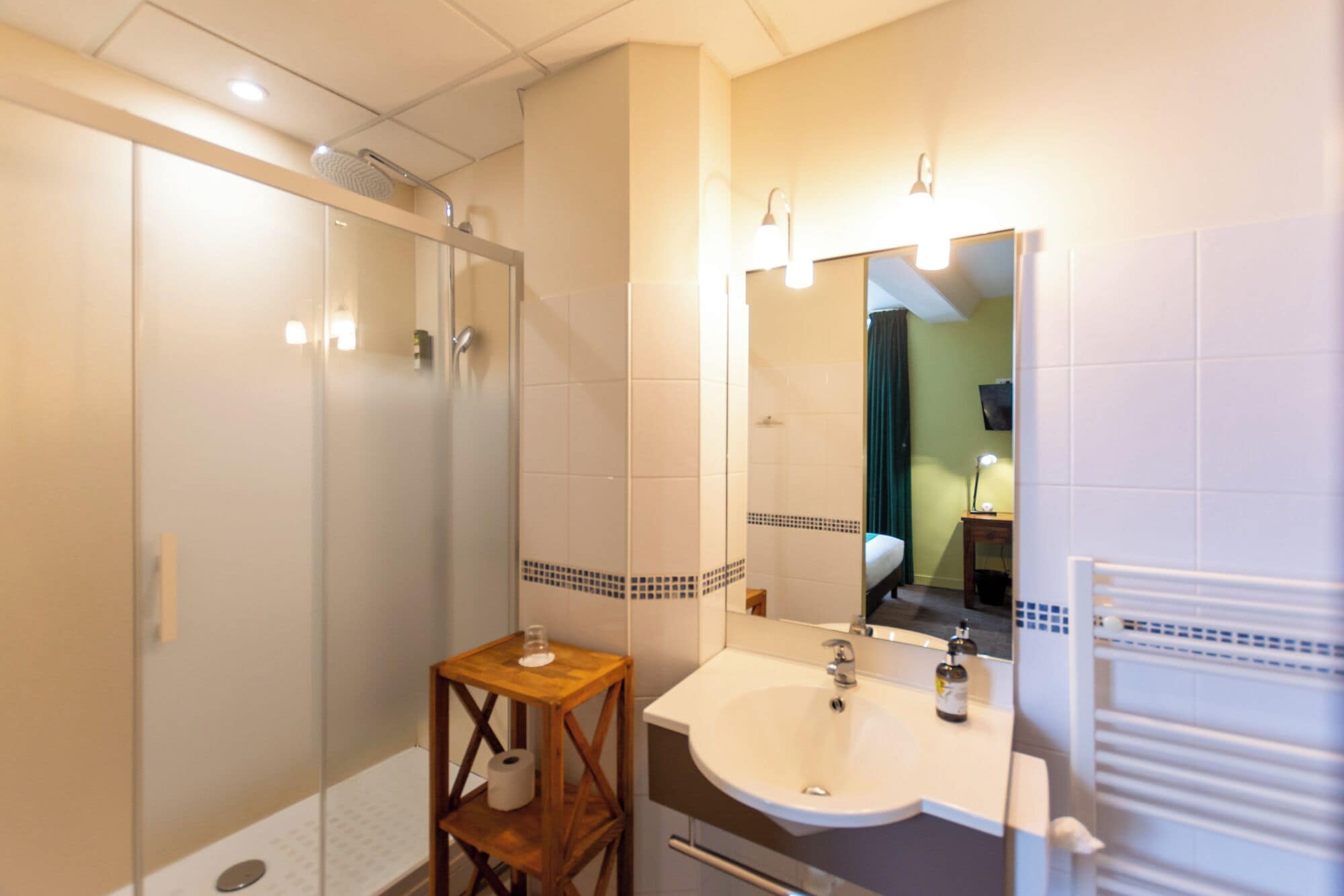 A bathroom with a glass shower enclosure, a wooden stool, a sink with a mirror, and a towel rack.