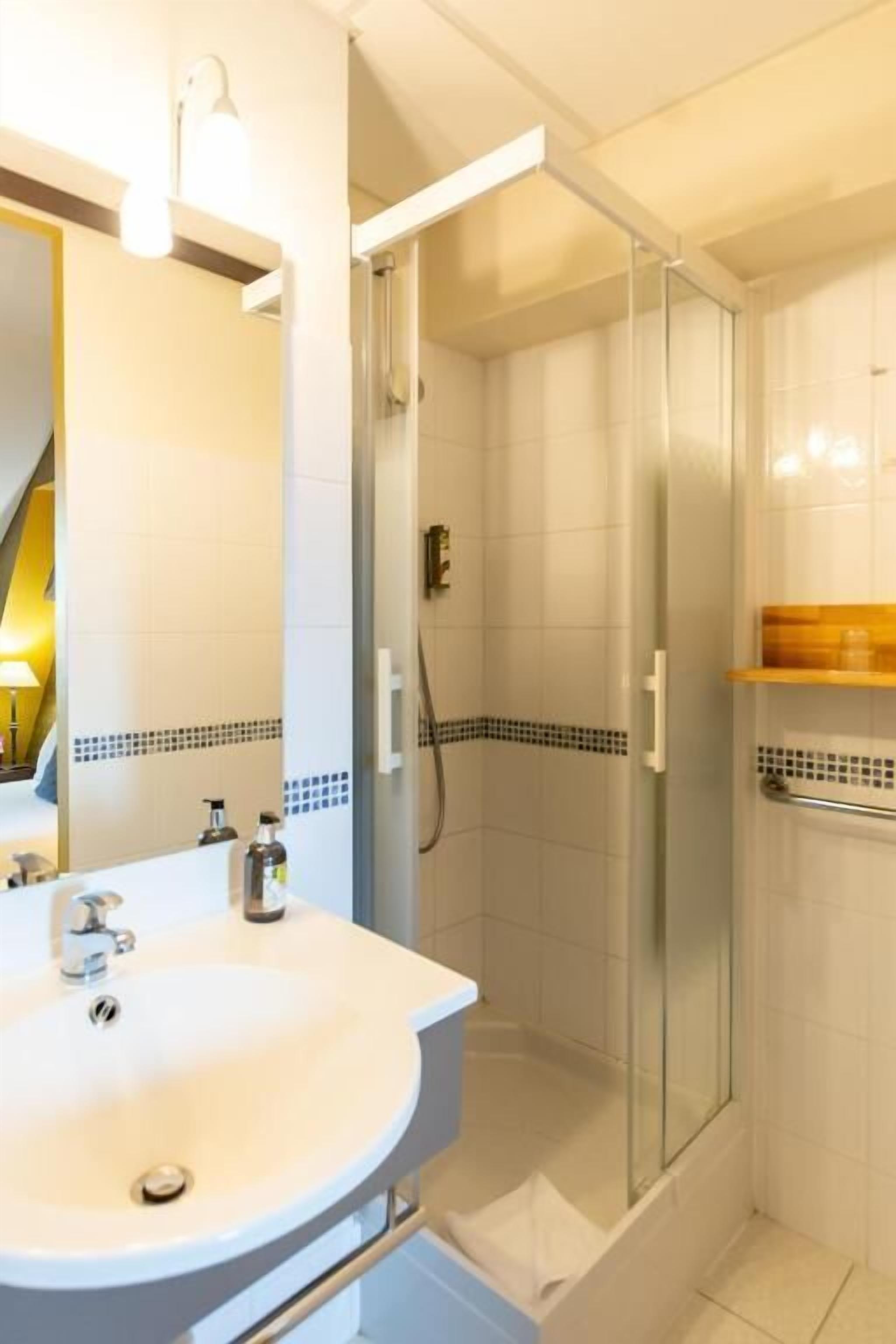 Un baño con lavamanos blanco, cabina de ducha con mampara de vidrio y estante de madera.