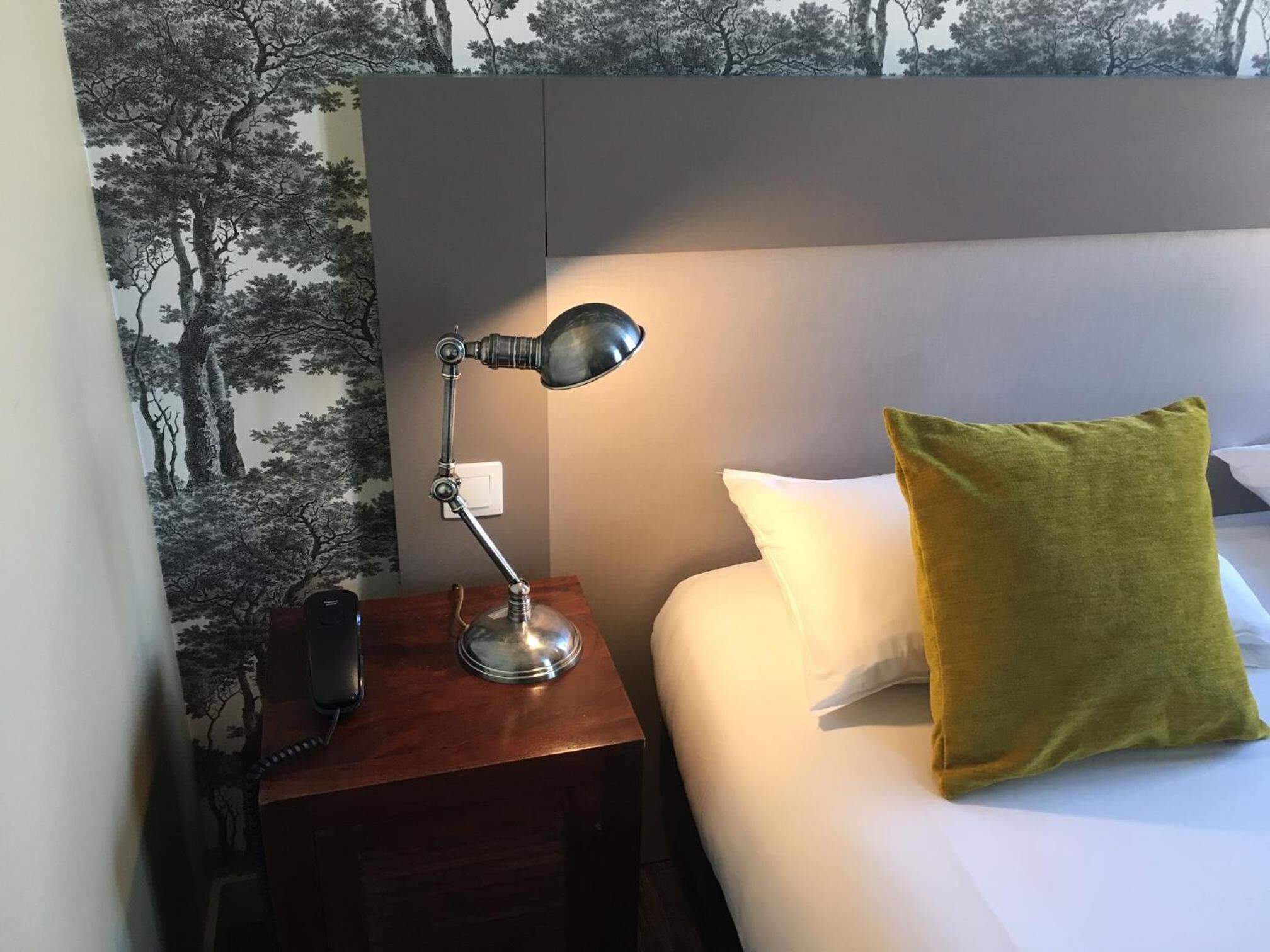 Un lit bien fait, avec un oreiller blanc et un oreiller de couleur vert moutarde, une table de chevet avec une lampe et un téléphone.